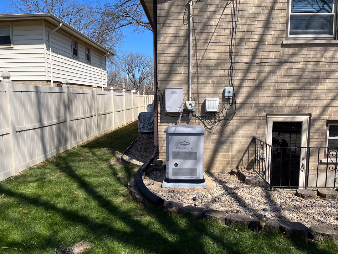 Briggs & Stratton backup generator installed on a gravel bed beside a brick house with utility meters and wiring above.