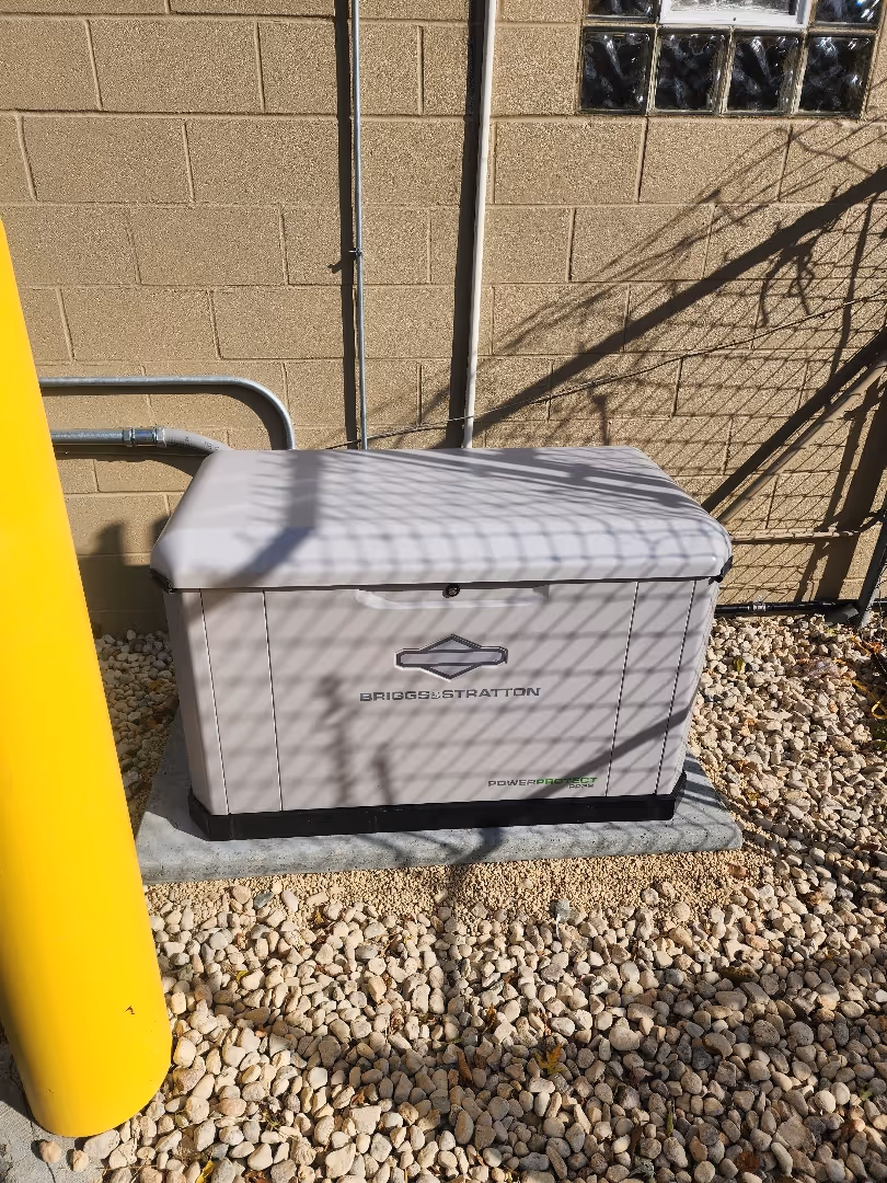 Briggs & Stratton outdoor generator installed on a concrete pad surrounded by gravel, with a yellow protective pole to the left.