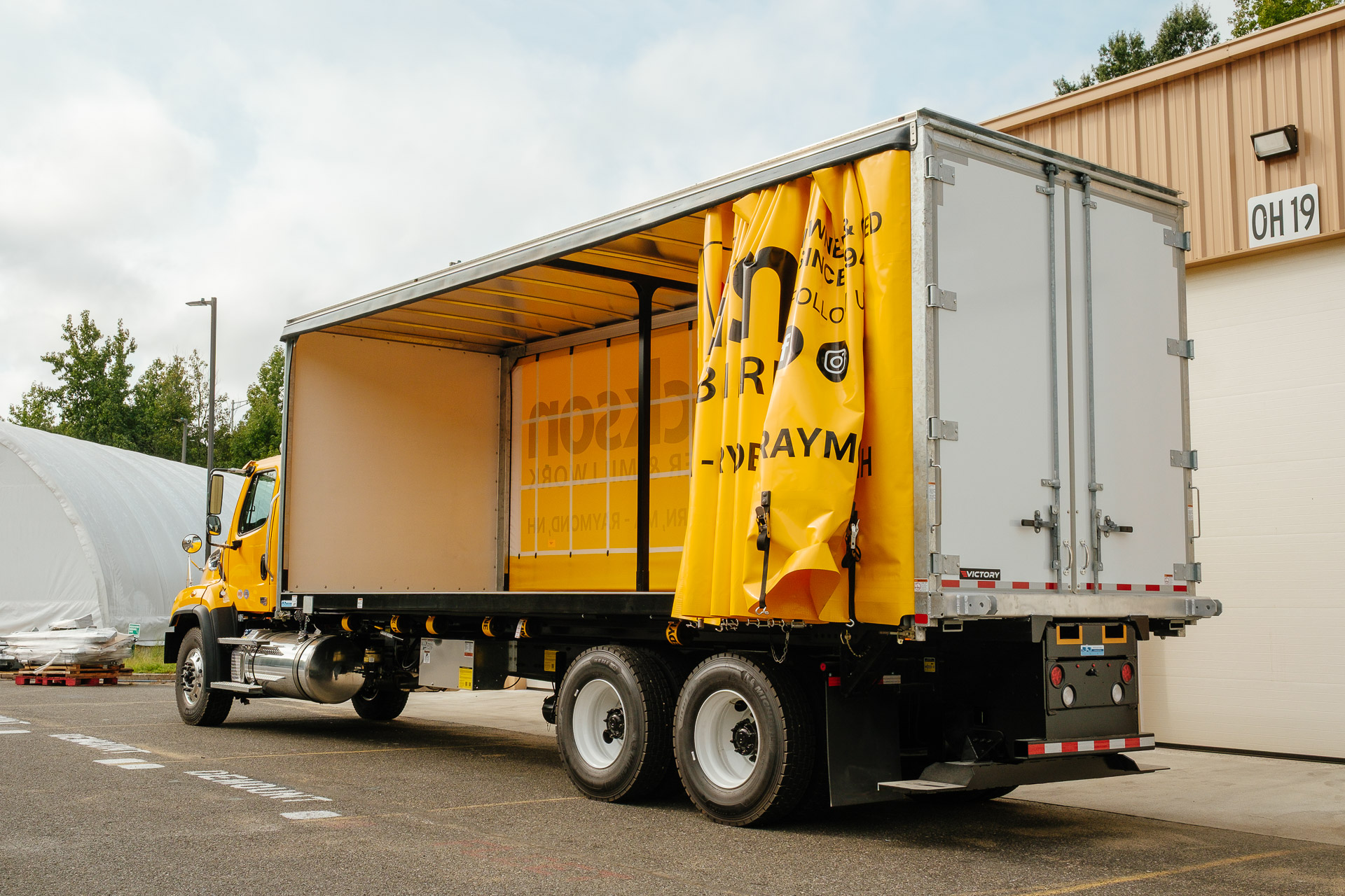A curtainside truck with it's yellow curtain open and parked in front of a warehouse.