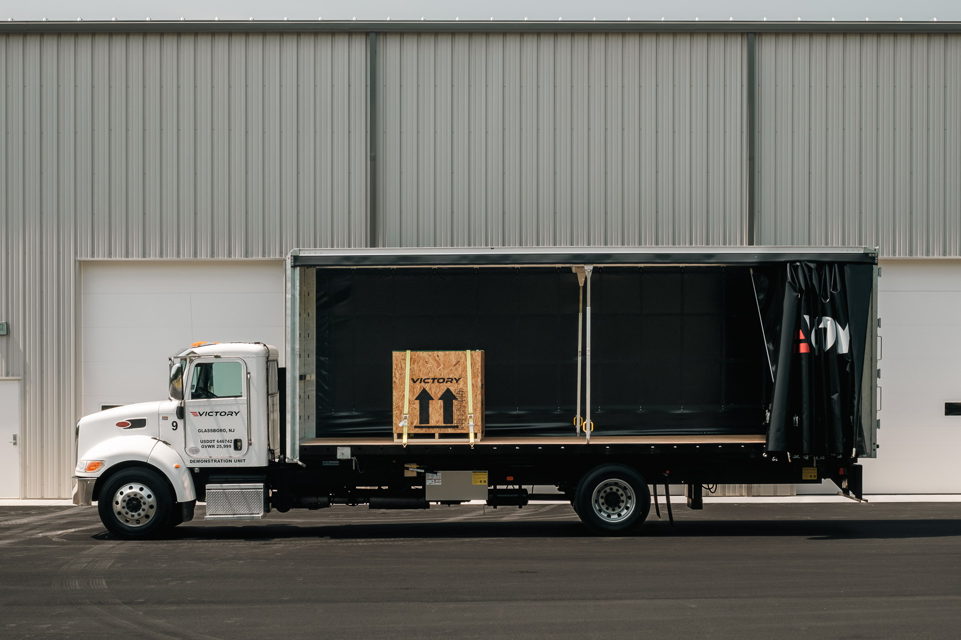side view of a curtainside truck with the curtain fully open, parked in front of a warehouse. A box of cargo is strapped down inside the truck's body.