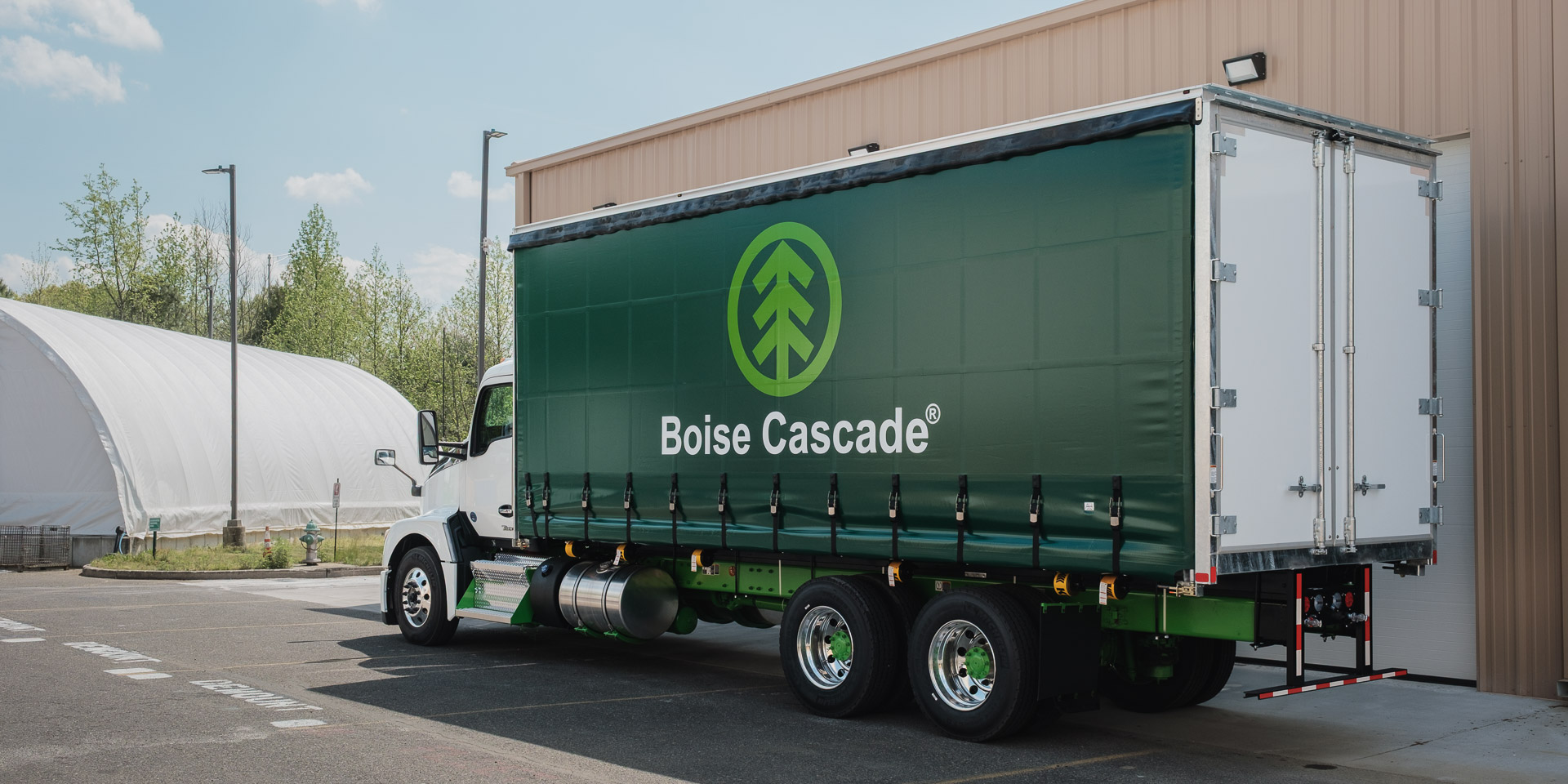 A green curtainside truck with the curtain closed and the logo for Boise Cascade on it is parked in front of a warehouse.