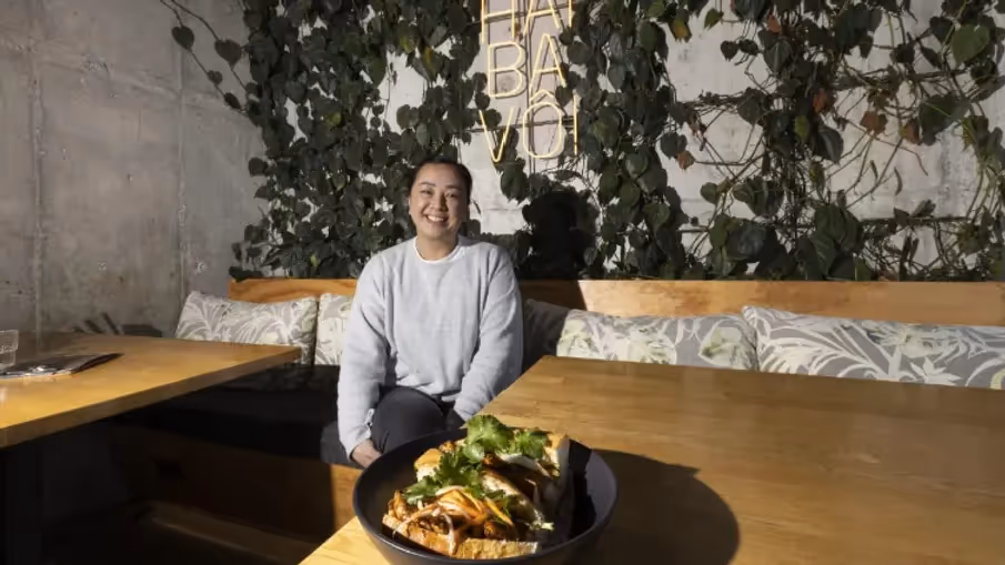 "Smiling woman sitting in a trendy Hamilton restaurant with lush greenery, showcasing a delicious plate of food. The inviting atmosphere highlights Hamilton's vibrant dining scene, supporting local businesses and community experiences."