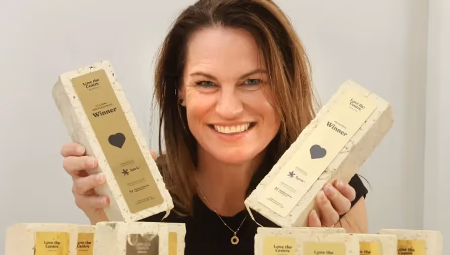 A smiling woman proudly holds up award-winning trophies made from local materials, surrounded by more trophies. Showcasing Hamilton's spirit, Love the Centre promotes community achievements and supports local businesses.