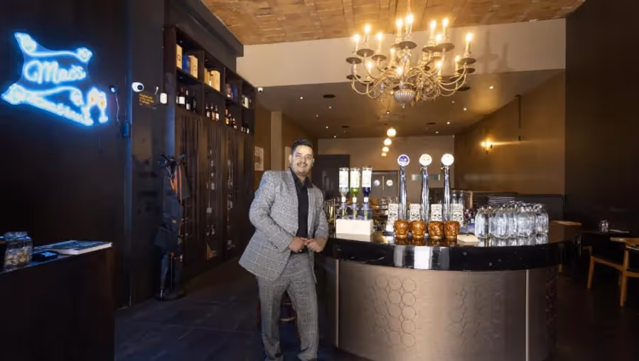 Alt text: "A smiling bartender in a stylish venue featuring a modern bar with taps and glasses. The space exudes elegance, with a chandelier and neon sign, highlighting Hamilton's vibrant nightlife and local dining scene."