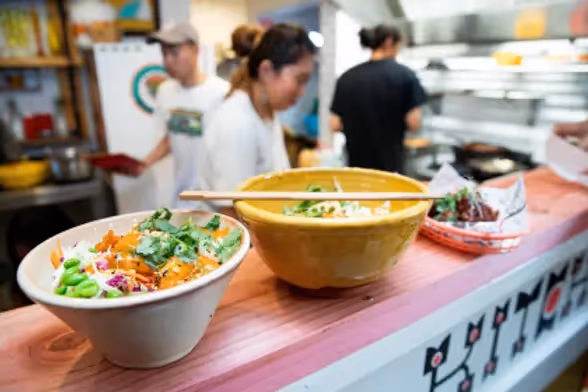 Alt text: Close-up of two vibrant bowls of fresh, locally-prepared salads on a wooden counter, highlighting Hamilton's culinary scene. In the background, chefs are seen creating delicious dishes, showcasing community dining experiences.