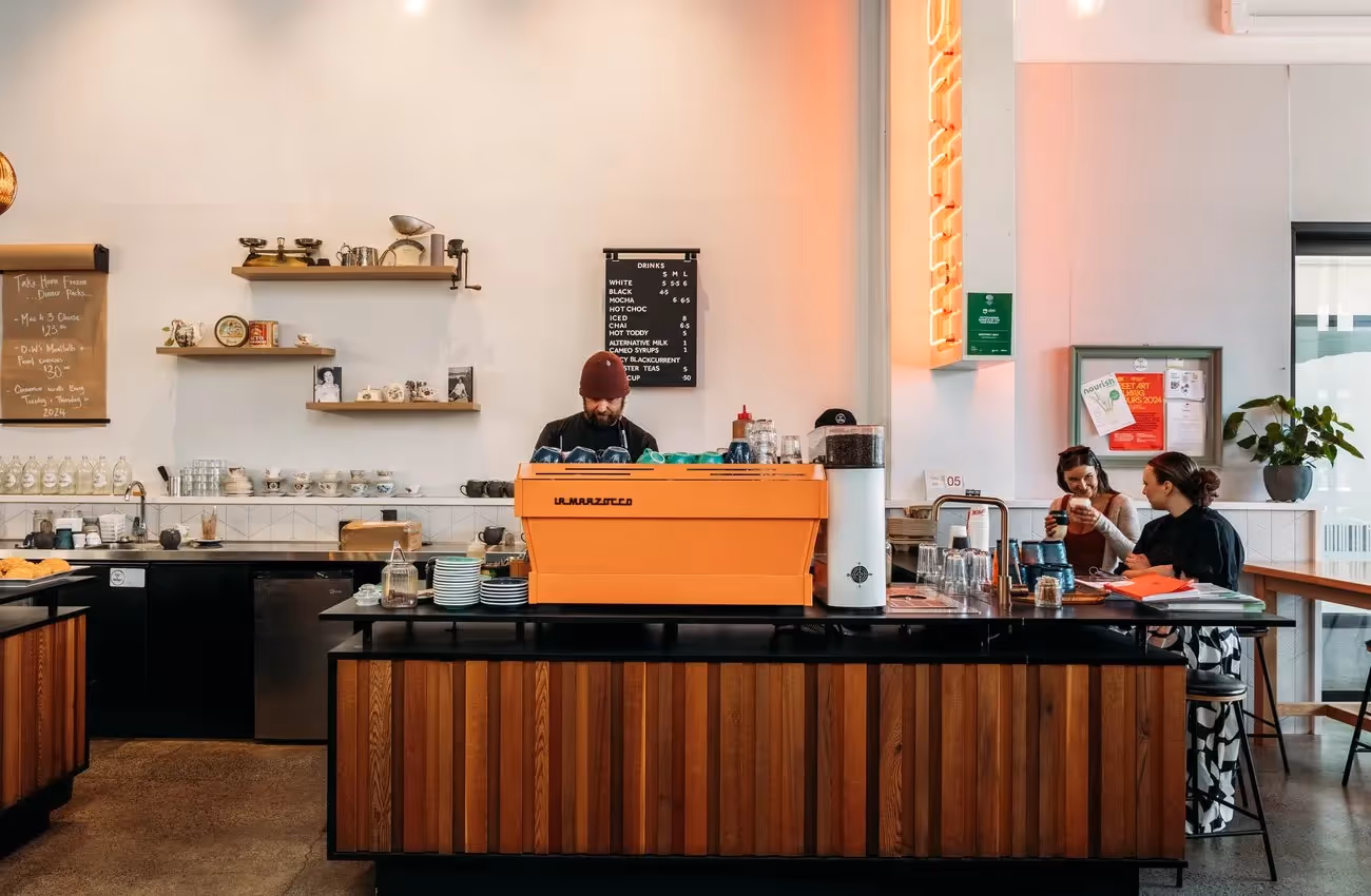 Alt text: "Barista preparing coffee at a vibrant Hamilton café, featuring sleek wooden counters and a lively atmosphere. Locals enjoy drinks, highlighting the community spirit and support for local businesses in Hamilton."
