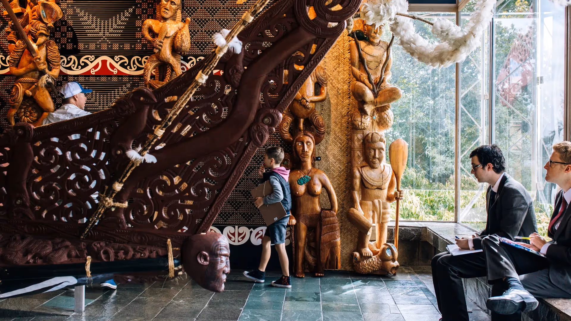 Alt text: "A young child walking through a vibrant cultural space in Hamilton, surrounded by intricate wooden carvings and traditional Māori art. Two men in suits observe the scene, highlighting the city's community and local heritage."