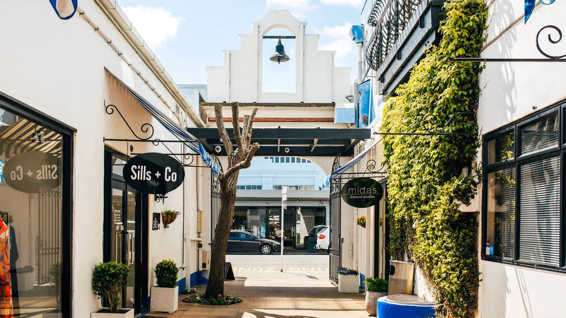 Alt text: "Charming shopping alley in Hamilton featuring local boutiques Sills + Co and Midas, framed by lush greenery and a distinctive architectural archway. A welcoming space that highlights Hamilton's vibrant community and local businesses."