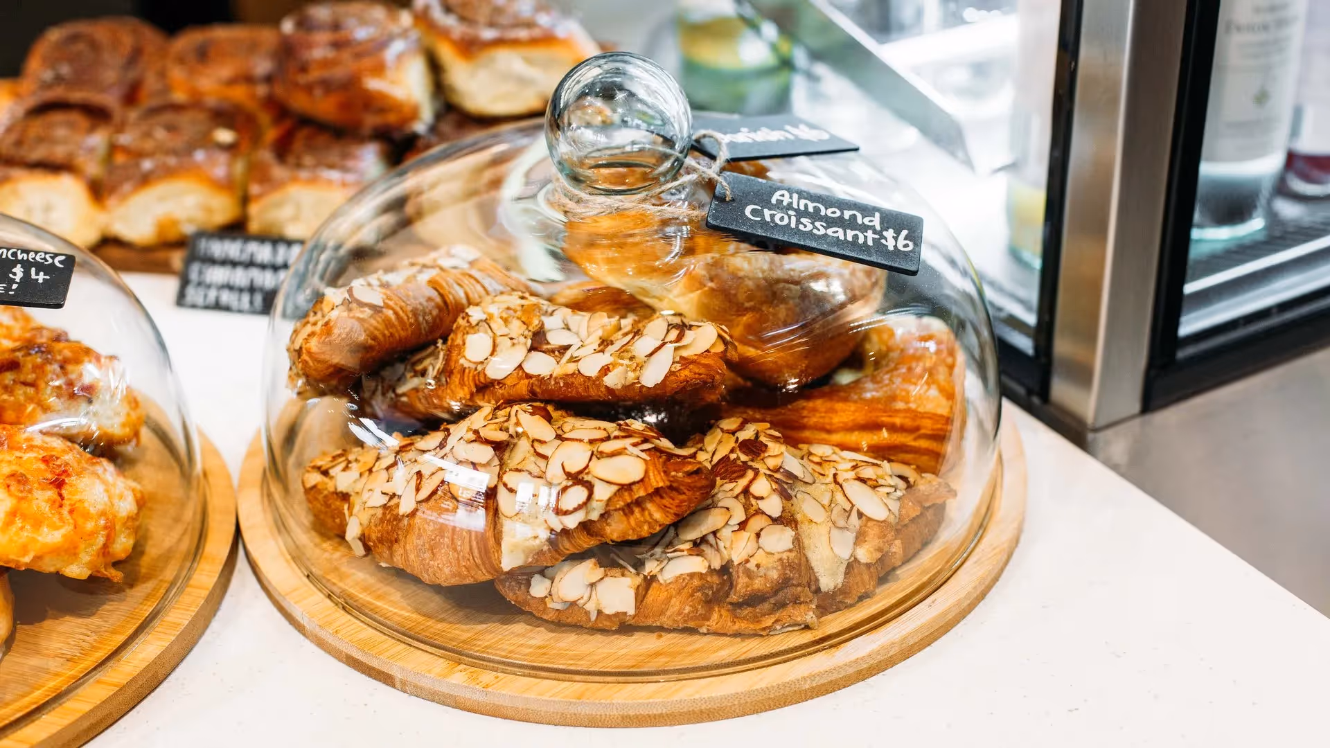Delicious almond croissants displayed under a glass dome on a wooden serving platter, highlighting local pastries in Hamilton. Perfect for bakery lovers seeking sweet treats in the heart of the city. Support local businesses with every bite.