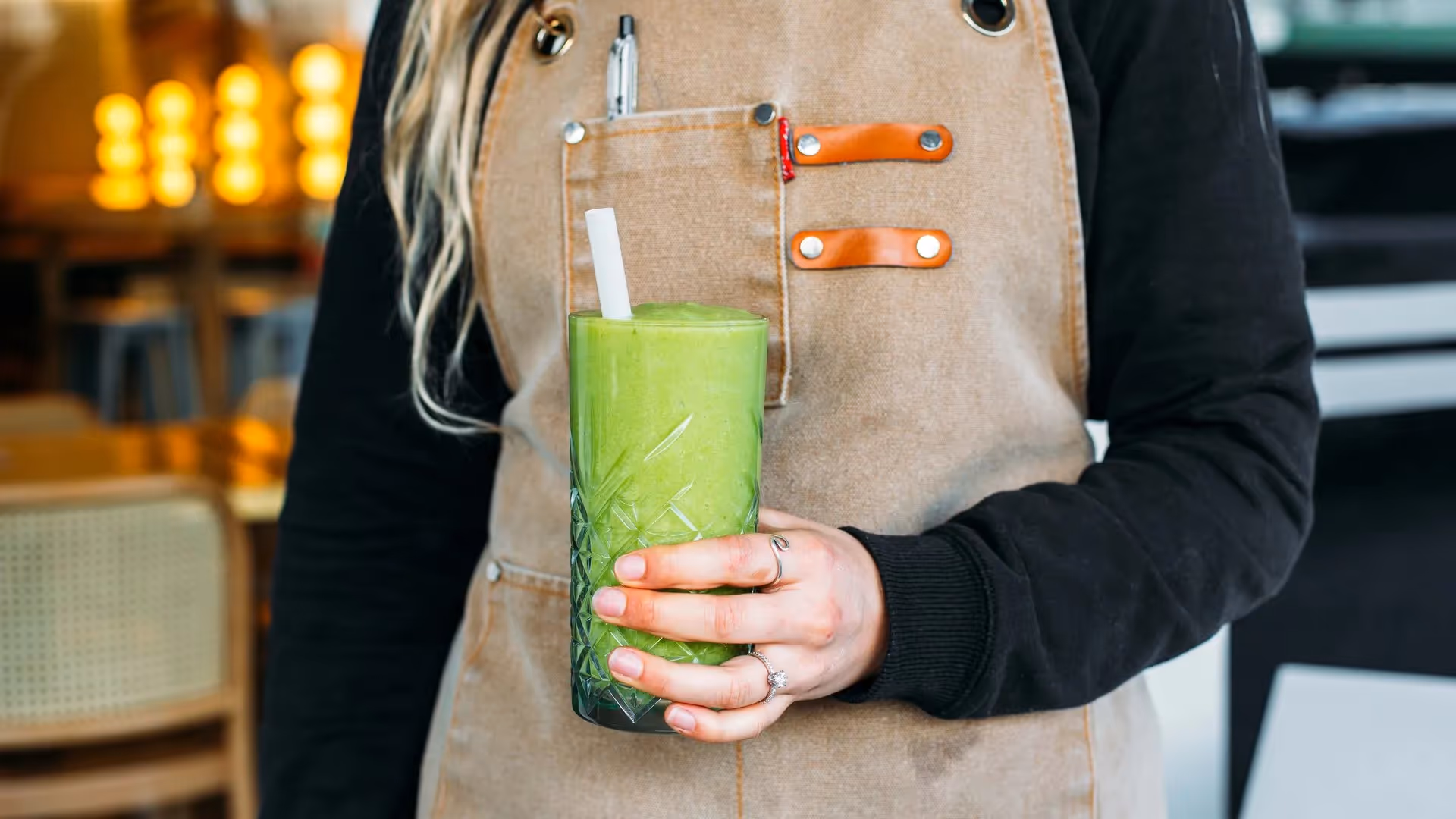 A barista in a tan apron holds a vibrant green smoothie in a decorative glass, showcasing Hamilton's café culture. This image highlights local food and drink experiences, promoting community engagement and supporting local businesses.