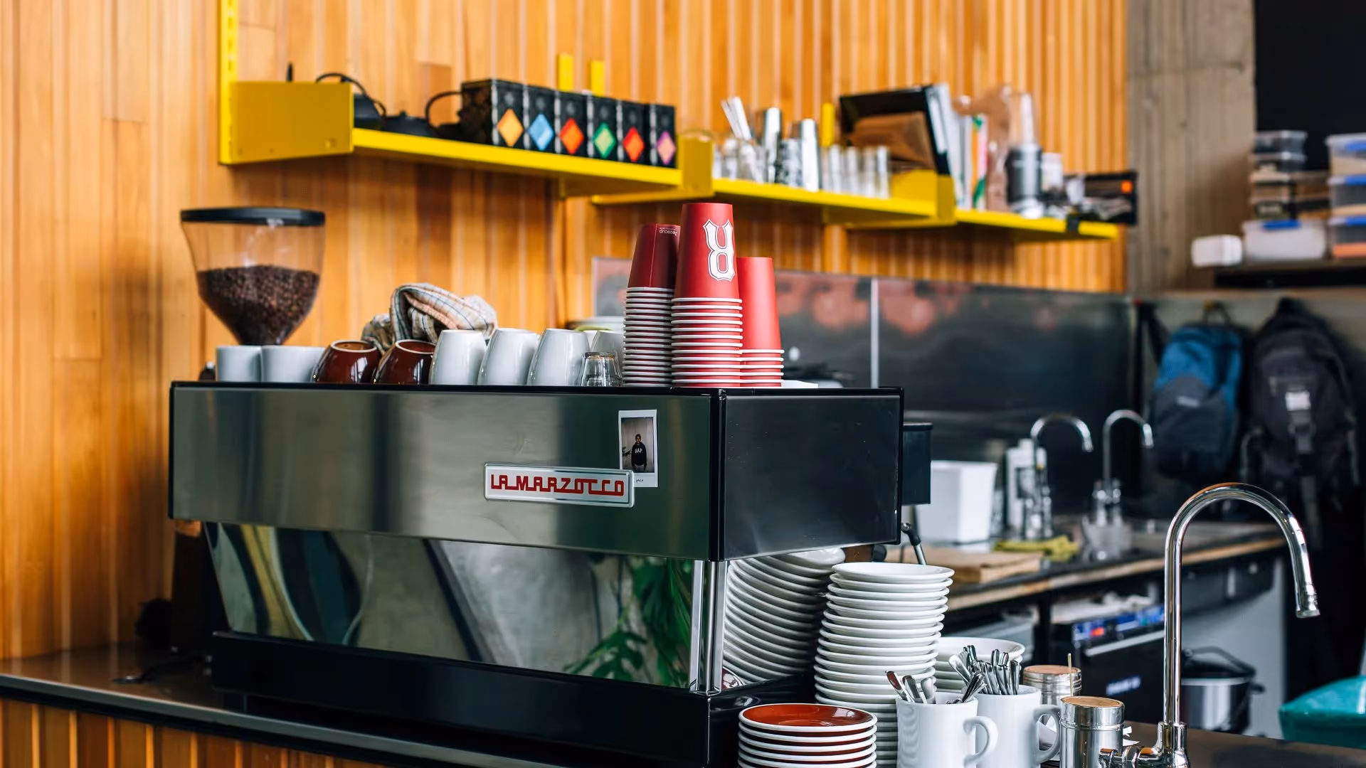 Alt text: A vibrant café scene in Hamilton featuring a La Marzocco espresso machine, stacked coffee cups, and a backdrop of wooden walls. This image highlights the city's local coffee culture, supporting community businesses and experiences.