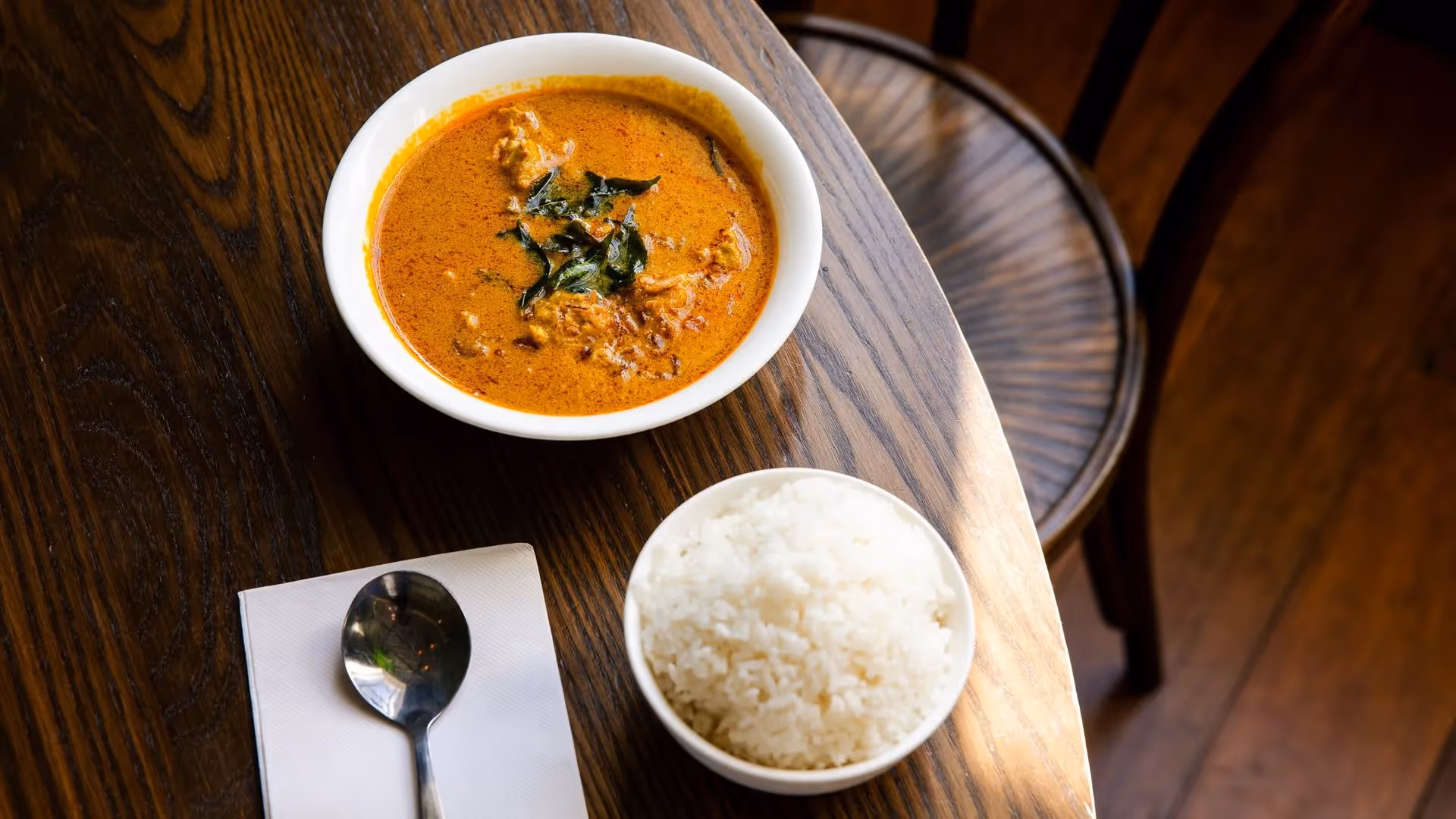 A bowl of rich curry topped with fresh herbs sits next to a serving of fluffy white rice, displayed on a rustic wooden table. This appetising dish highlights Hamilton's vibrant food scene and local culinary delights.