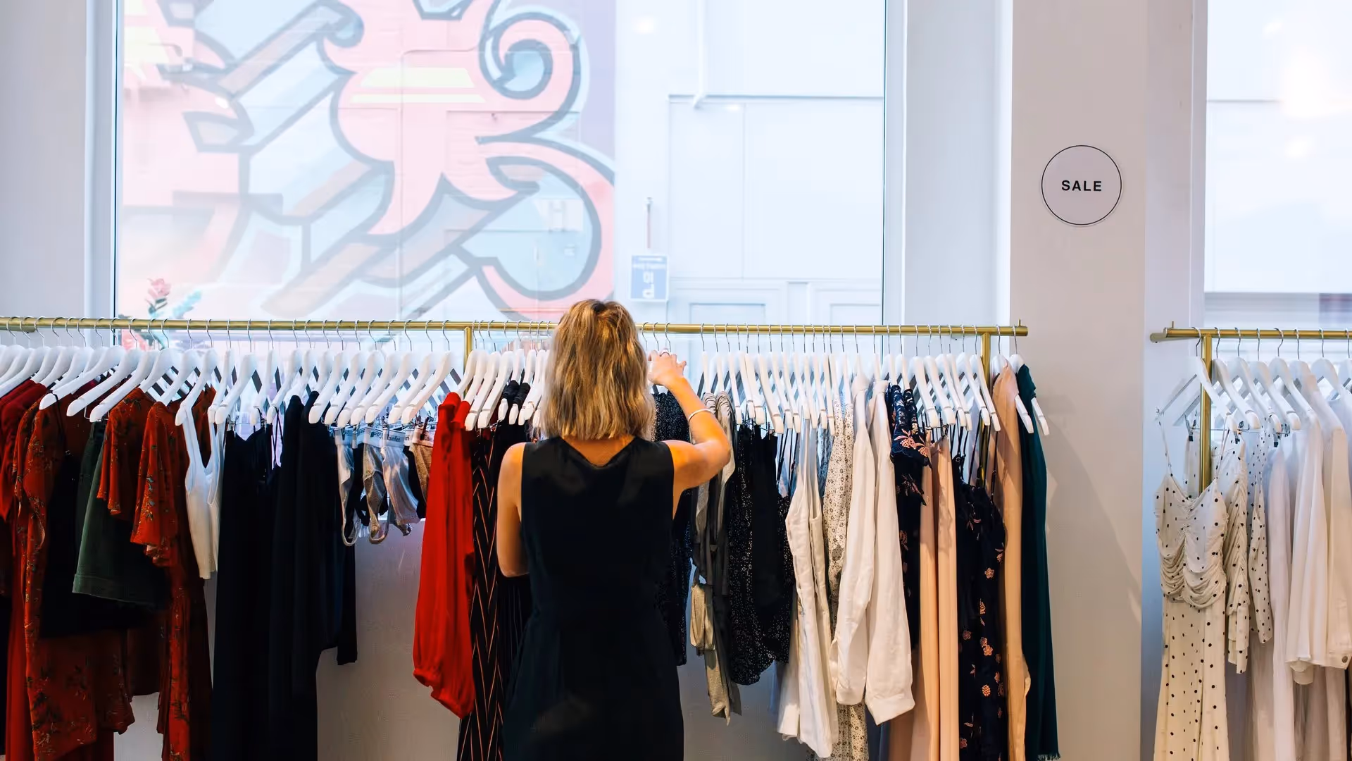 Alt text: A shopper browsing a vibrant clothing rack in a Hamilton boutique, surrounded by a variety of stylish garments on sale. The shop's decor features artistic murals, highlighting local creativity and supporting community businesses.