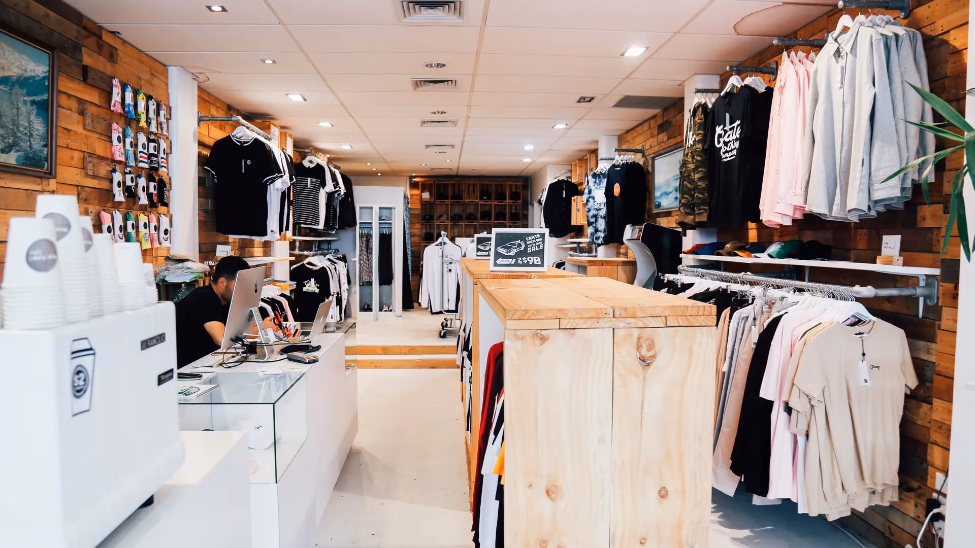 Alt text: Interior of a trendy retail shop in Hamilton showcasing clothing and accessories. The bright space features wooden decor, neatly arranged apparel, and a friendly staff member at the checkout, promoting local business and community spirit.