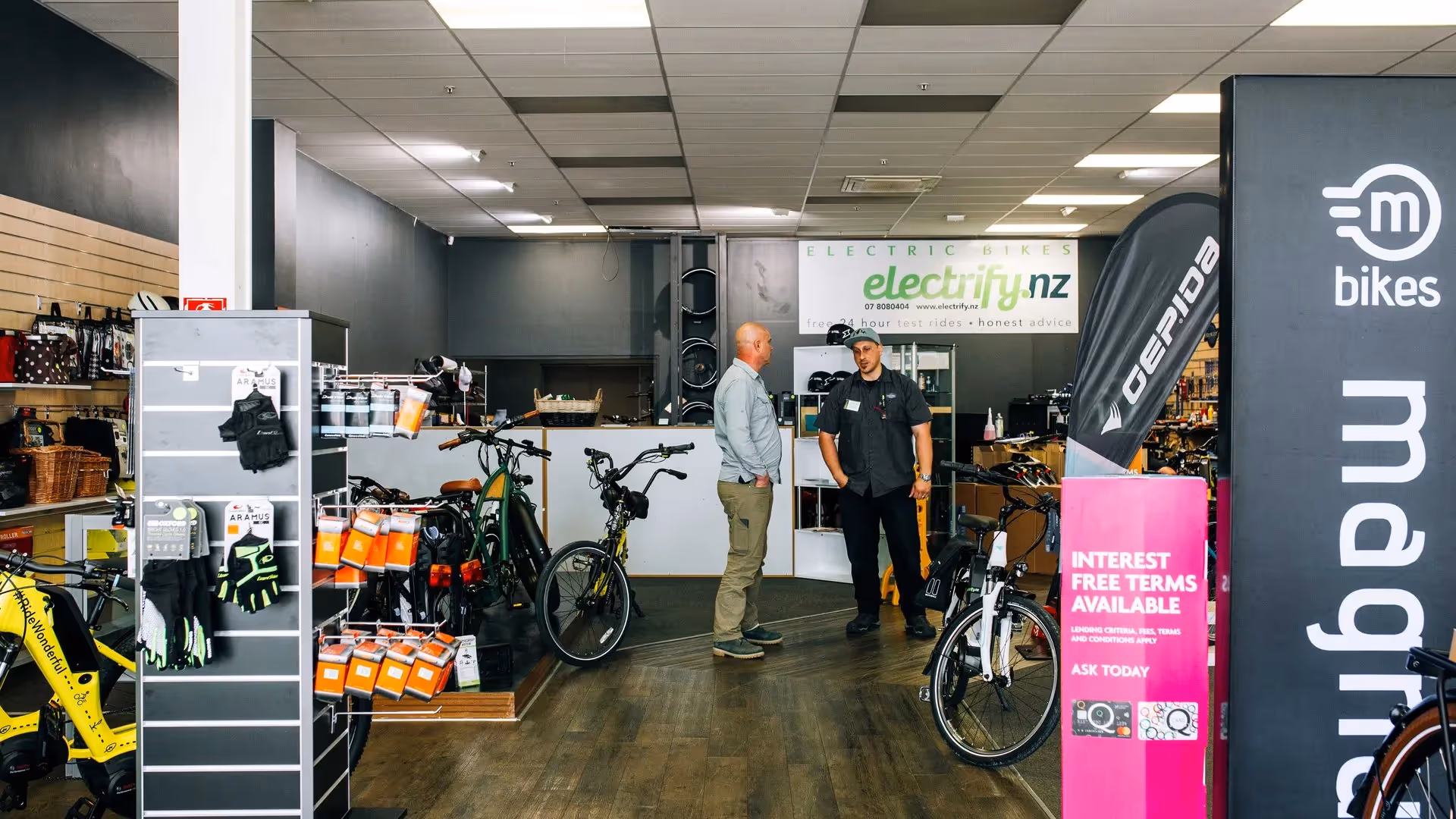 Alt text: Two men discussing electric bikes in a modern bike shop featuring a range of bicycles and accessories. The store promotes local businesses in Hamilton, showcasing innovative e-bike options and community-focused experiences.