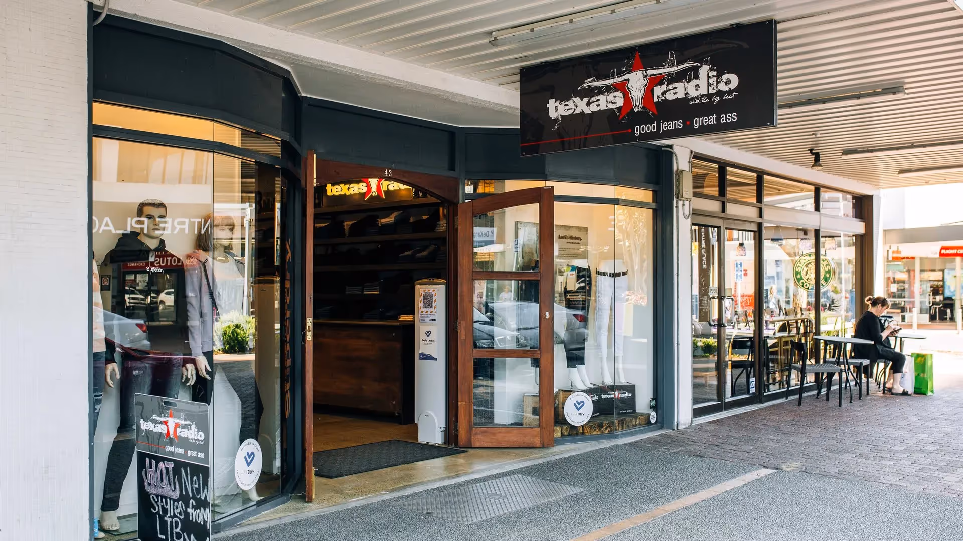 Alt text: "Exterior view of Texas Radio store in Hamilton, showcasing a welcoming entrance and signage. A customer enjoys a coffee nearby, highlighting local community life and support for Hamilton's businesses."