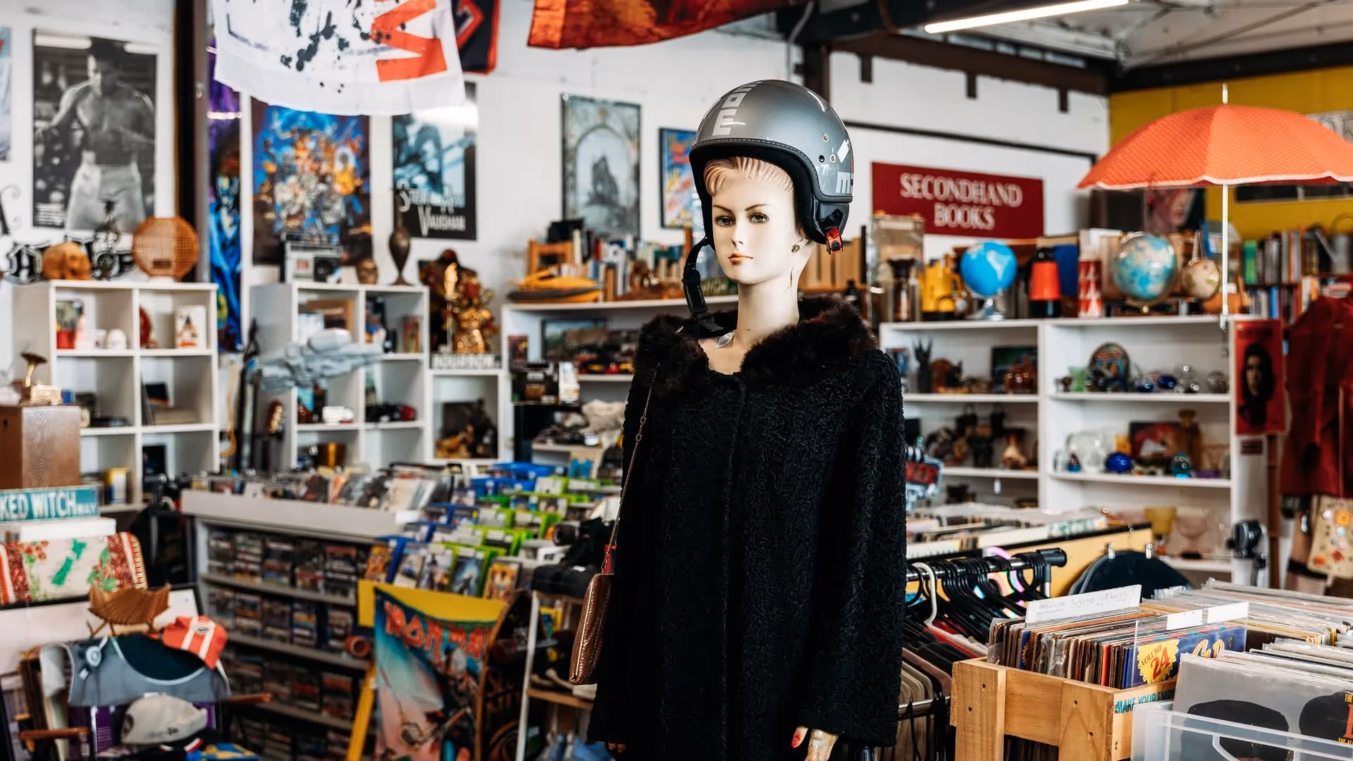 A vintage mannequin wearing a grey helmet stands in a vibrant second-hand store filled with eclectic items, including books, records, and local memorabilia. This scene highlights Hamilton's unique shopping experience and community spirit.