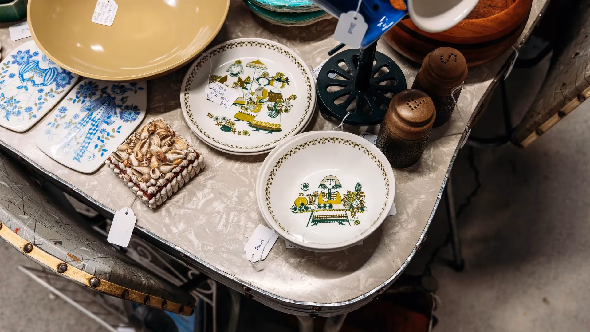 Alt text: A collection of vintage plates featuring charming illustrations, alongside decorative items and cutlery on a rustic table. This scene showcases Hamilton's local markets, celebrating community heritage and supporting local artisans.