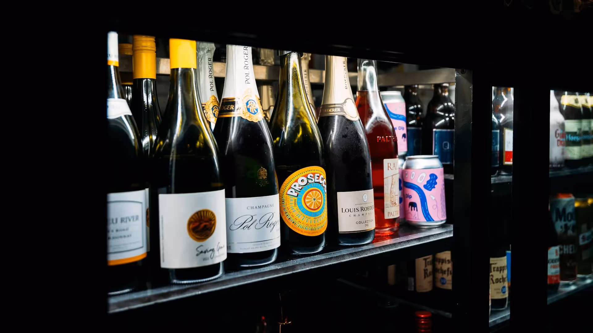 Alt text: A selection of fine wines and sparkling beverages displayed on a shelf, featuring bottles of Prosecco, Louis Brut, and various local drinks. Perfect for showcasing Hamilton's vibrant local businesses and community events in the heart of the city.