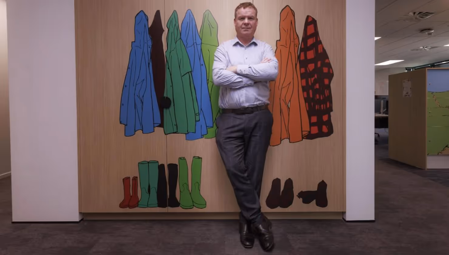 Alt text: A man stands confidently with crossed arms in front of a colourful mural depicting various coats, boots, and shoes. This image reflects the community spirit of Hamilton, showcasing local art and support for local businesses.