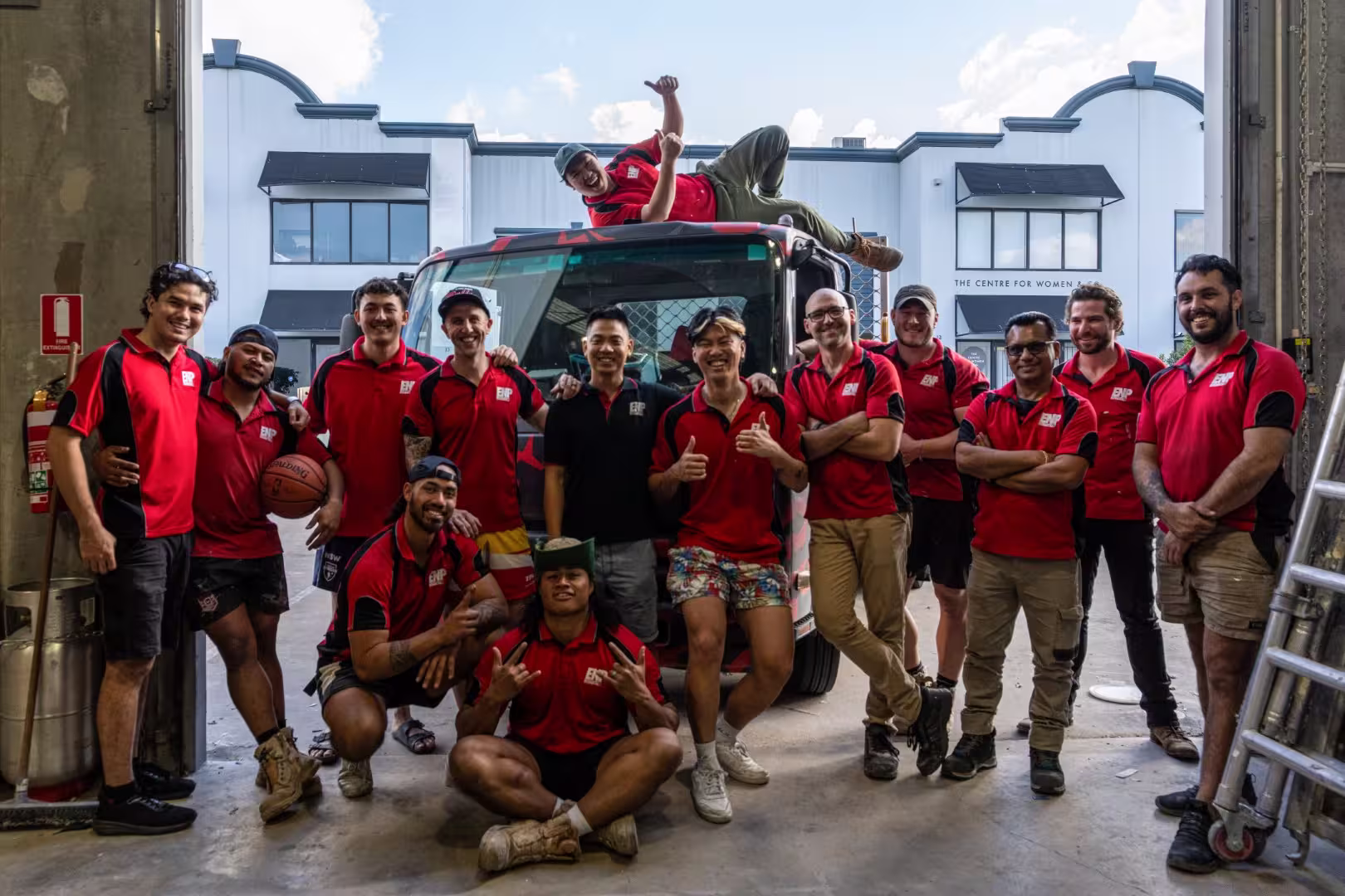 Group of men in red and black shirts posing playfully in front of a truck inside a warehouse.