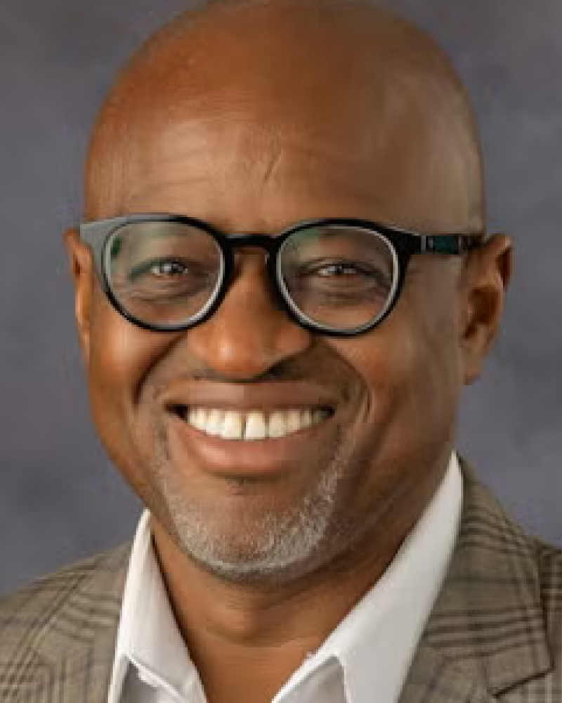 Smiling bald man with glasses wearing a checkered jacket and white shirt against a gray background.