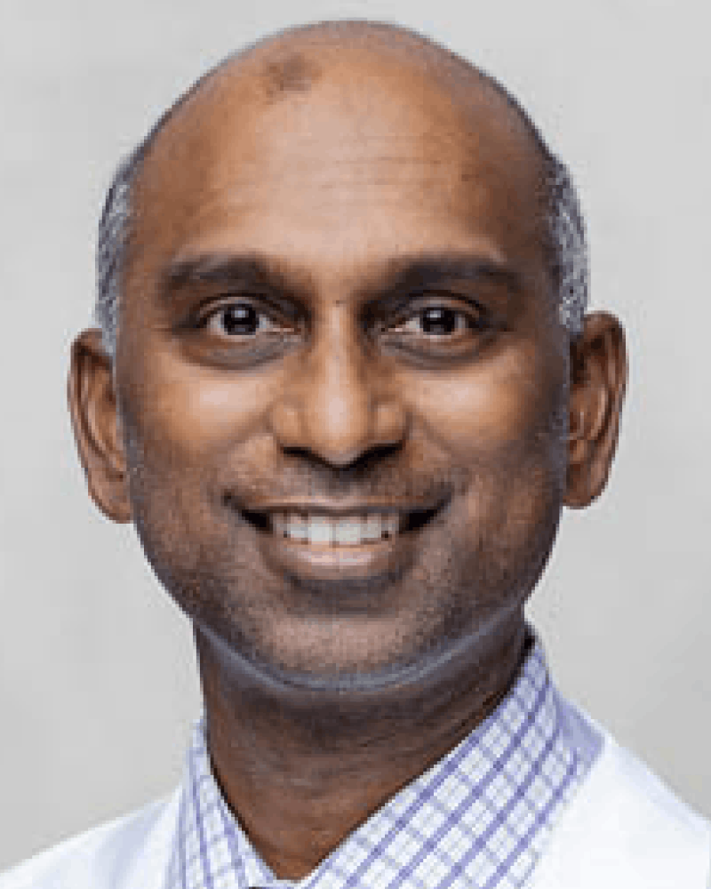 Close-up portrait of a smiling middle-aged man with dark skin, a bald head, and wearing a checkered shirt under a white coat.