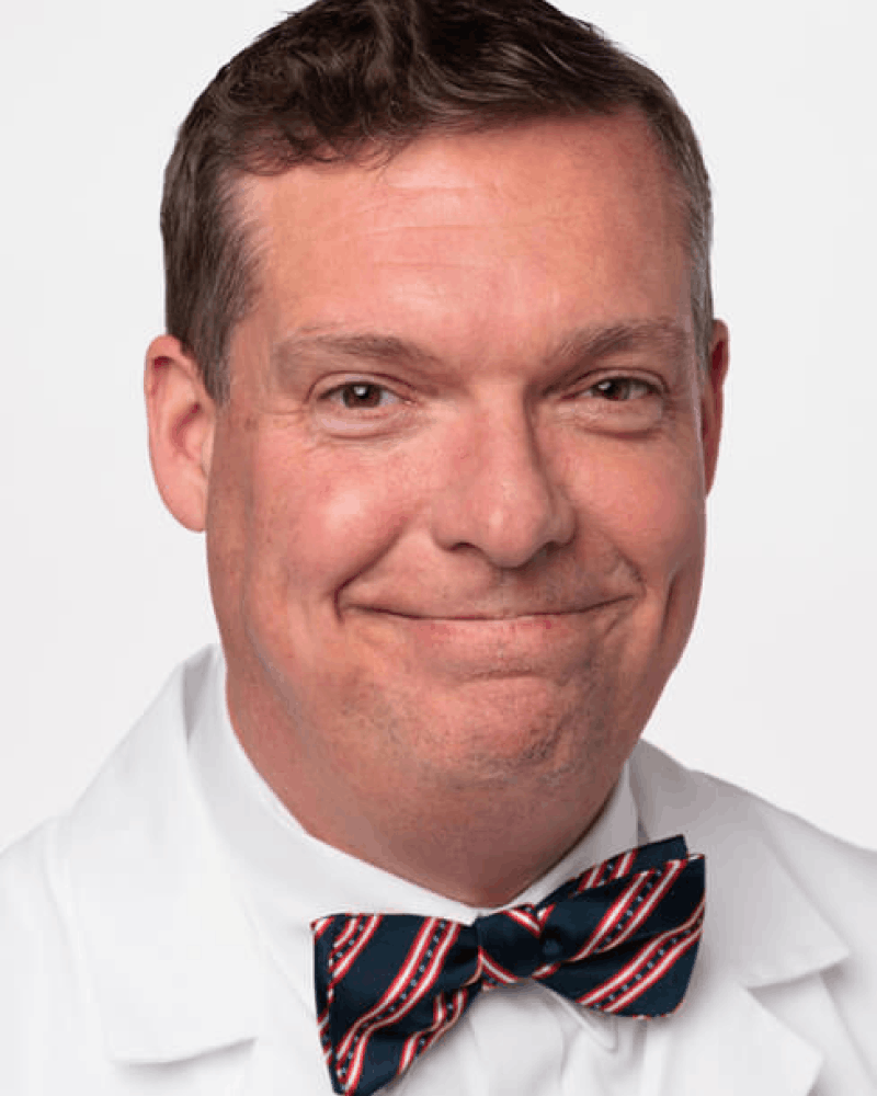 Smiling man with short brown hair wearing a white coat and a blue bow tie with red and white diagonal stripes.