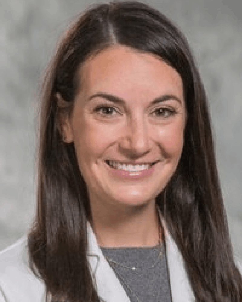 Smiling woman with long dark brown hair wearing a white coat over a gray top, set against a blurred gray background.
