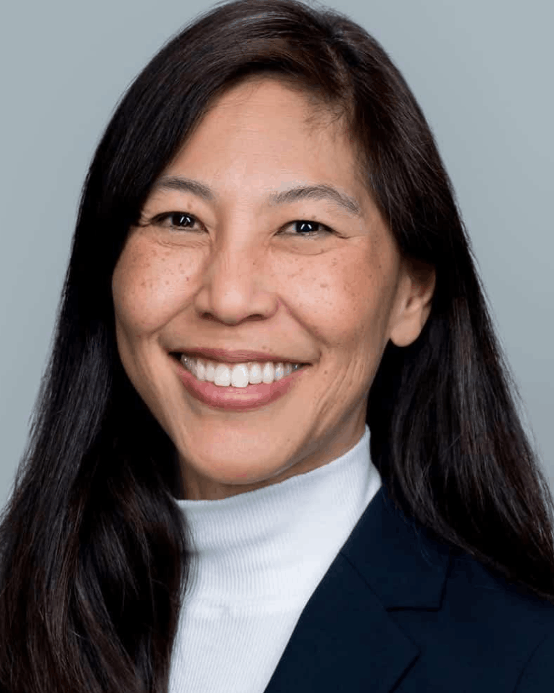 Smiling woman with long dark hair wearing a white turtleneck and dark blazer against a light gray background.