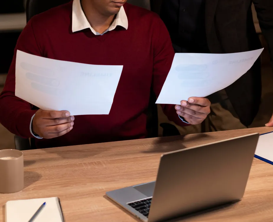 A man in a desk, holding 2 papers in front of laptop.
