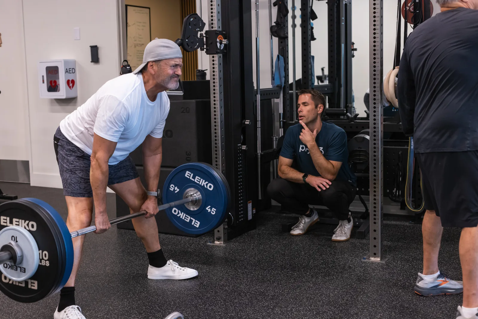 A proof3 coach watching a member in his fifties set up a barbell deadlift