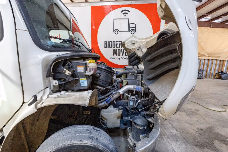 Open hood of a medium-duty truck in repair shop, exposing engine components, coolant reservoir, and hoses beside moving company trailer.