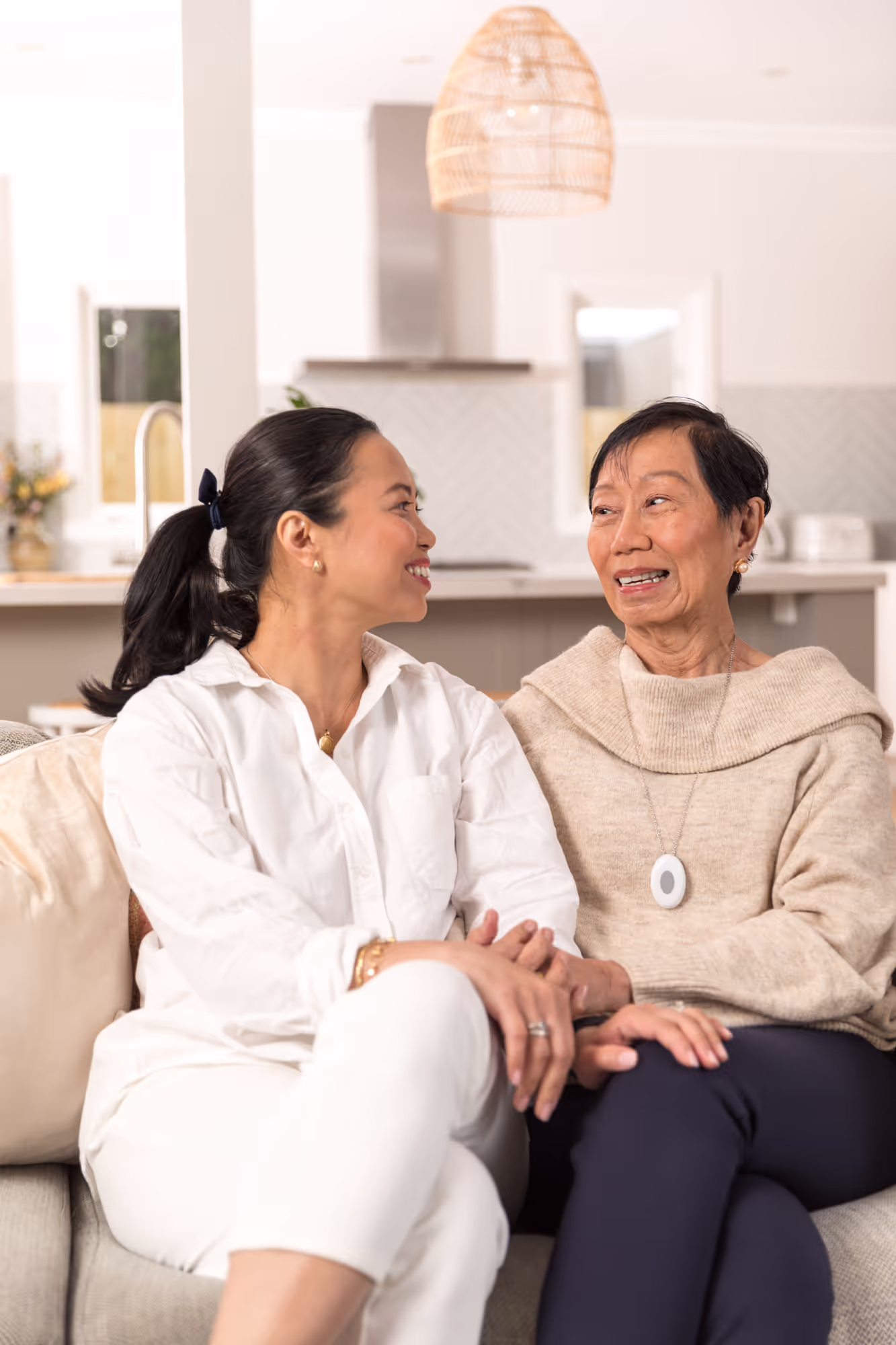 A mother and daughter sitting on the sofa and looking at each other. The mother is wearing a small falls detection personal alarm pendant around her neck.
