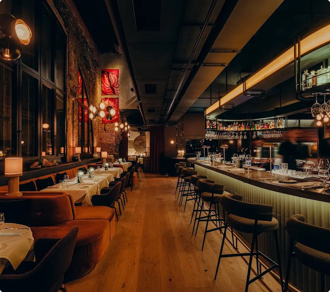 Warmly lit modern restaurant interior with a row of tables set with white tablecloths on the left and a bar with stools on the right.