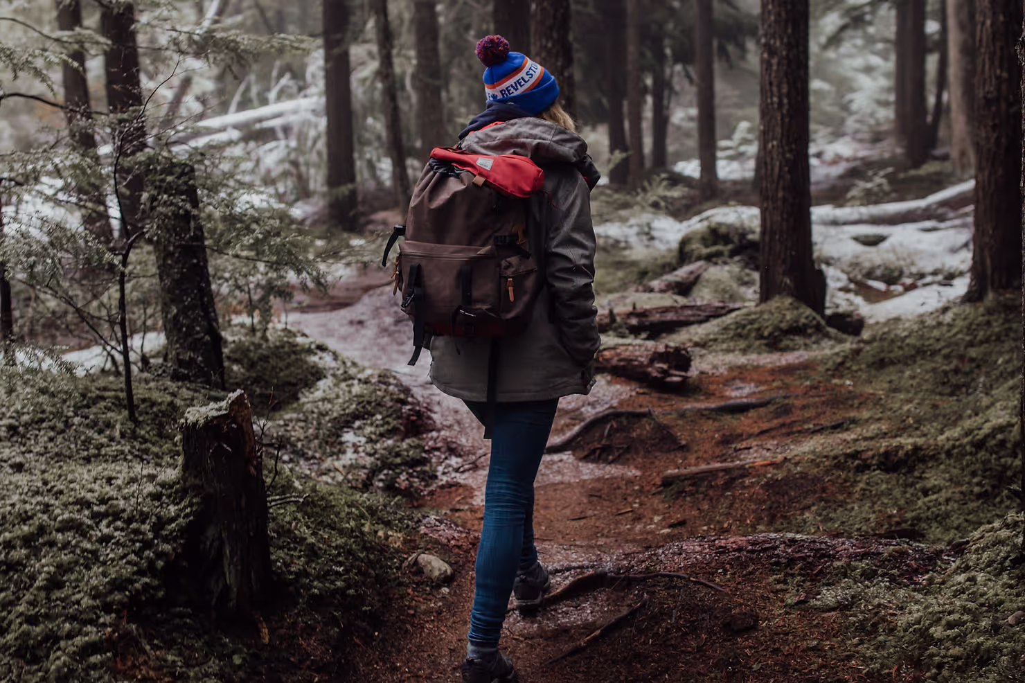 Hiker carrying a large backpack walks along a forest trail. The forest is dense with snow-dusted trees and moss-covered ground, creating a serene, wintry atmosphere.