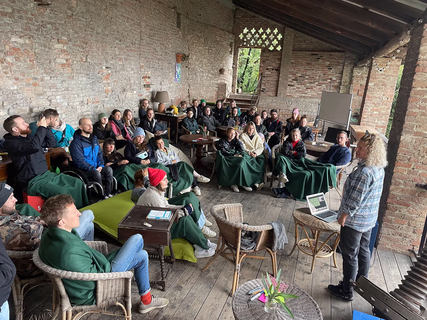 A person stands presenting with a laptop to a group of attentive people seated on wicker chairs, covered in green blankets, in a rustic, open-brick setting with wooden floors.