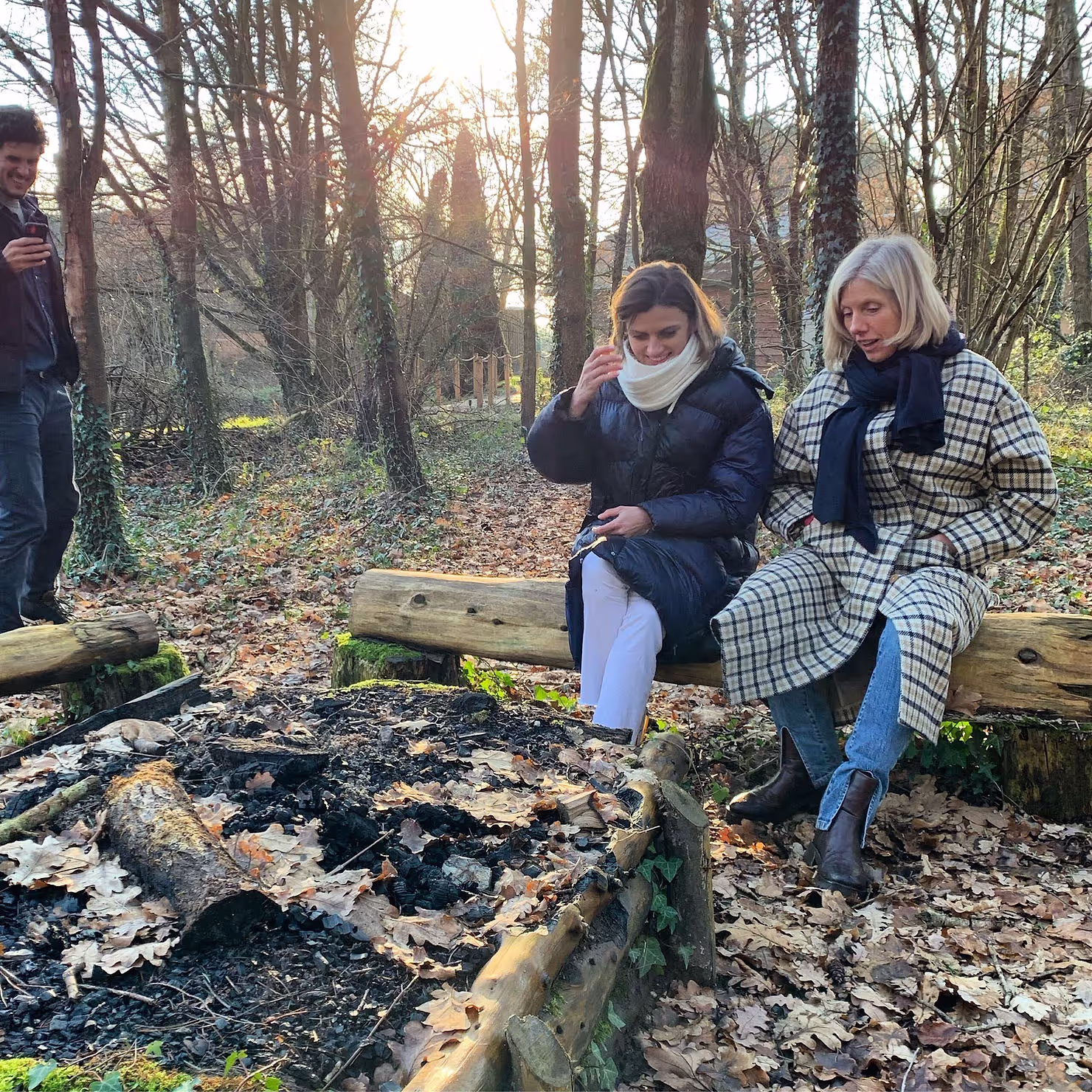 Two people sit on a log bench, wrapped in coats, next to an extinguished campfire in a leaf-strewn, sunlit forest clearing. A standing person observes them, holding a phone.