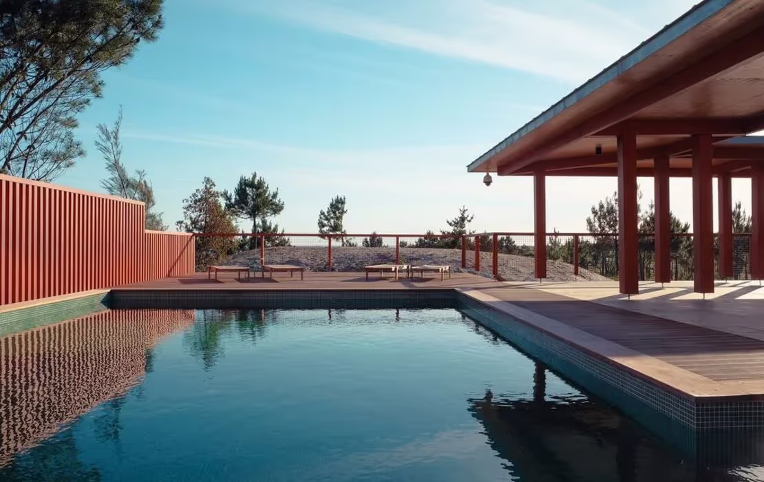 A swimming pool reflects the sky, bordered by a wooden deck and red fence. In the background, trees rise against a clear blue sky, creating a serene outdoor setting.