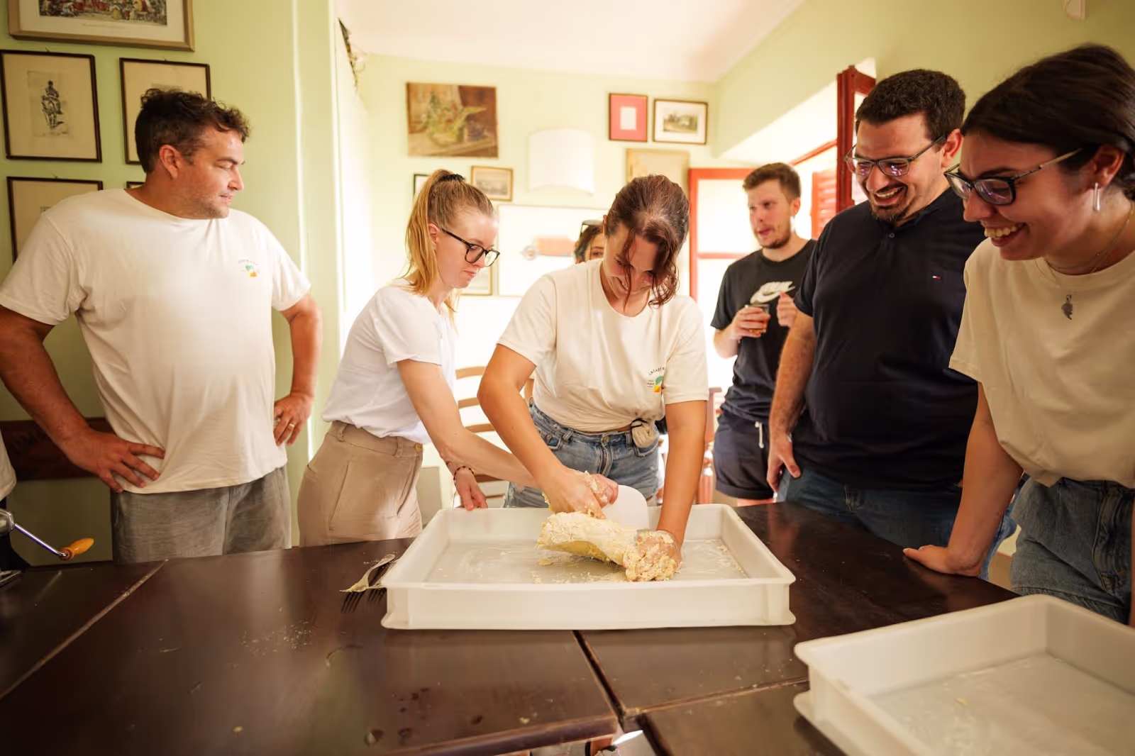 People gather around a table, smiling, as one person kneads dough in a large rectangular tray. The room has framed artworks on pale green walls.