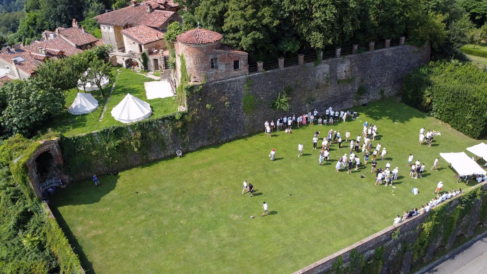 People gather in groups on a spacious green lawn, surrounded by historic buildings and tall stone walls, with trees and tents nearby, creating a lively outdoor event atmosphere.