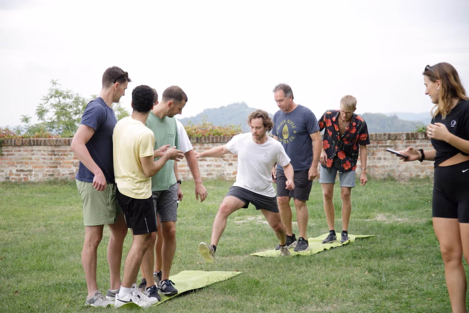 A group of six people participates in an outdoor exercise activity on grass, with one man jumping between mats while a woman holds a phone. A brick wall and hills are visible in the background.