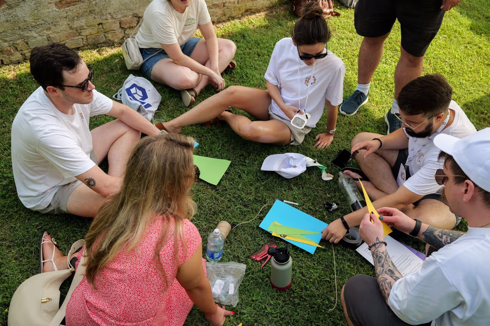 A group of people sit on grass, engaging in a collaborative activity involving colorful paper and tools. They are outdoors near a brick wall, appearing relaxed and focused.