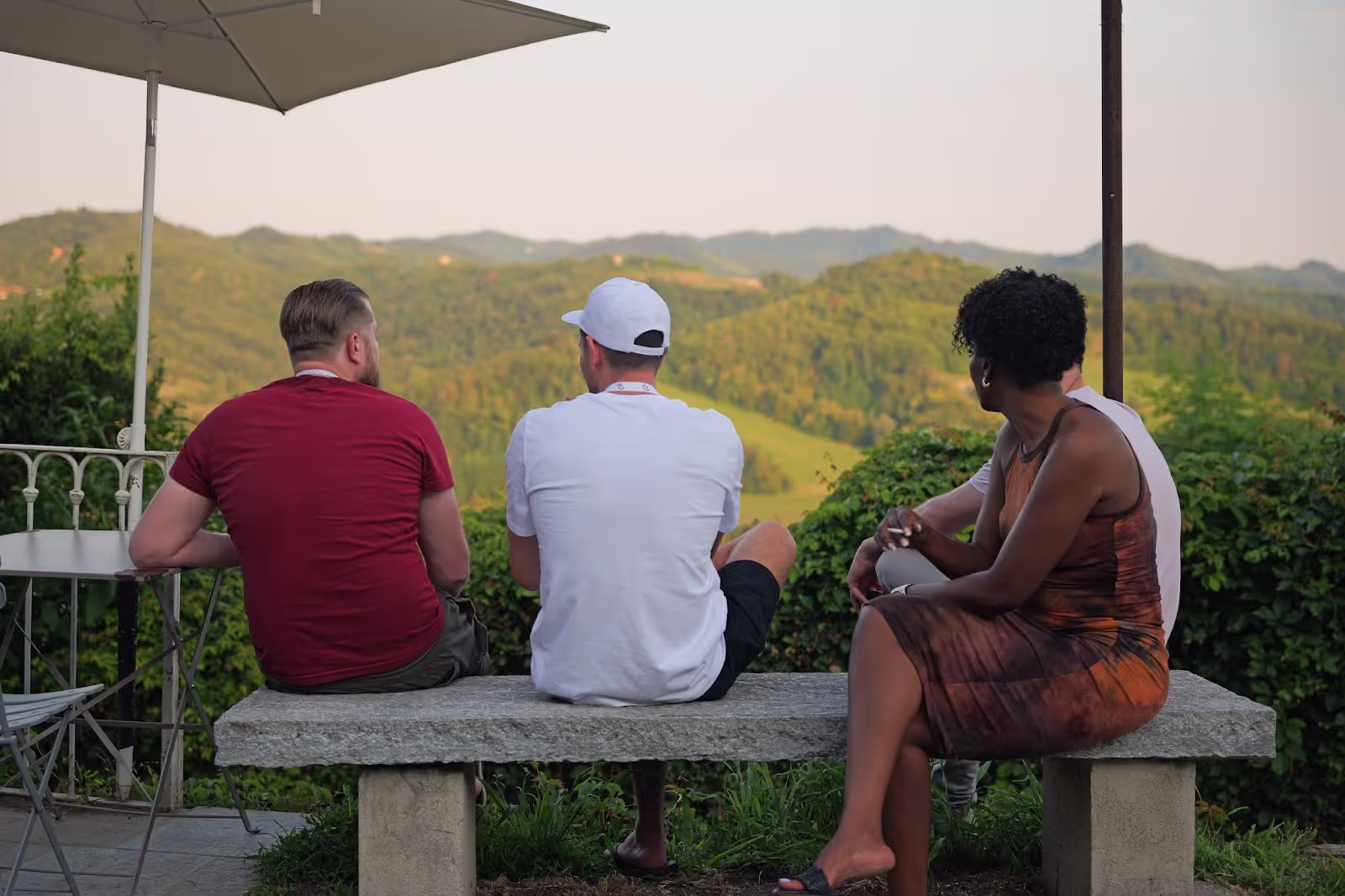 Three people sit on a stone bench under an umbrella, overlooking rolling green hills and distant mountains. A metal table and chair are nearby, surrounded by greenery.