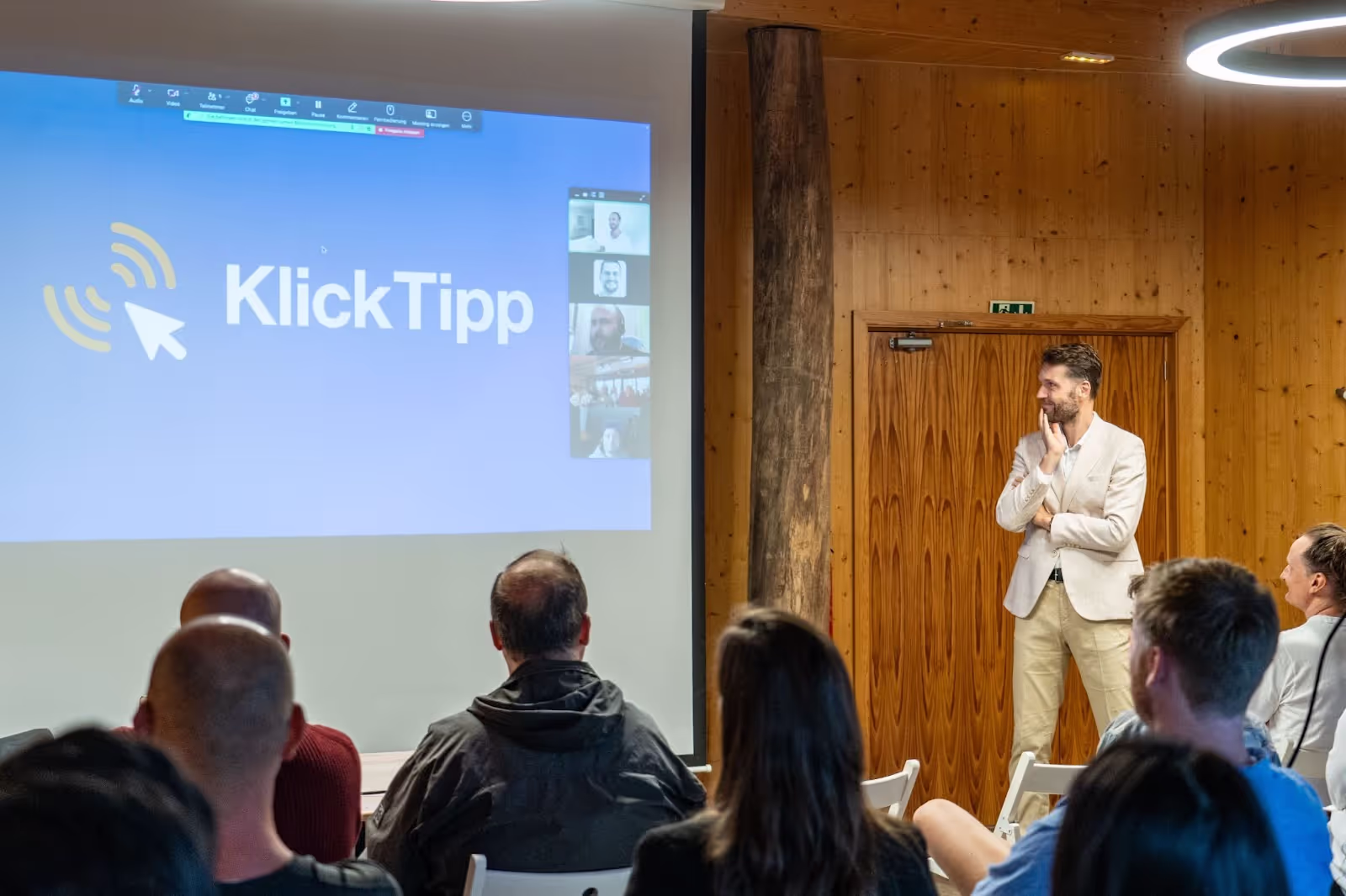 Presentation screen showing "KlickTipp" logo, with a person standing and observing, surrounded by seated audience in a wood-paneled room. Video call windows appear alongside the logo.
