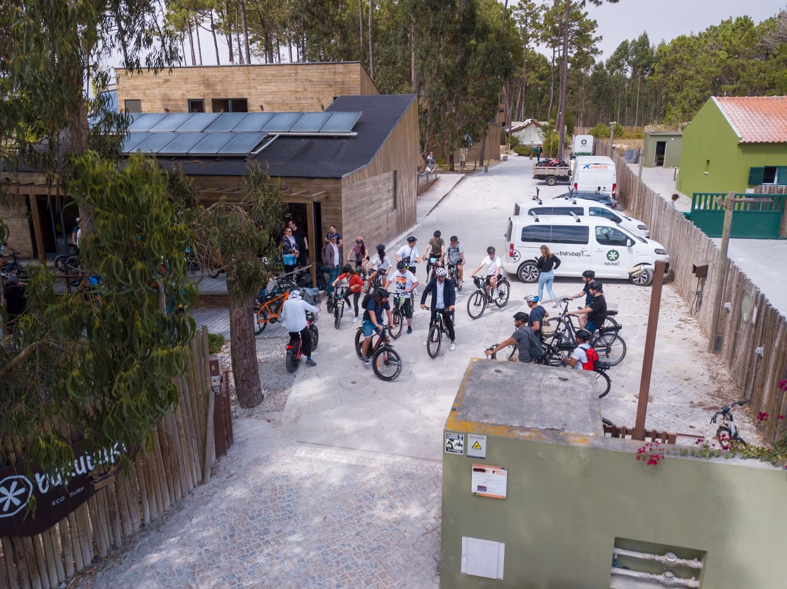 Cyclists gather outside a wooden building surrounded by trees, preparing for a ride. Nearby, two white vans with "bugaebike eco surf" branding are parked.