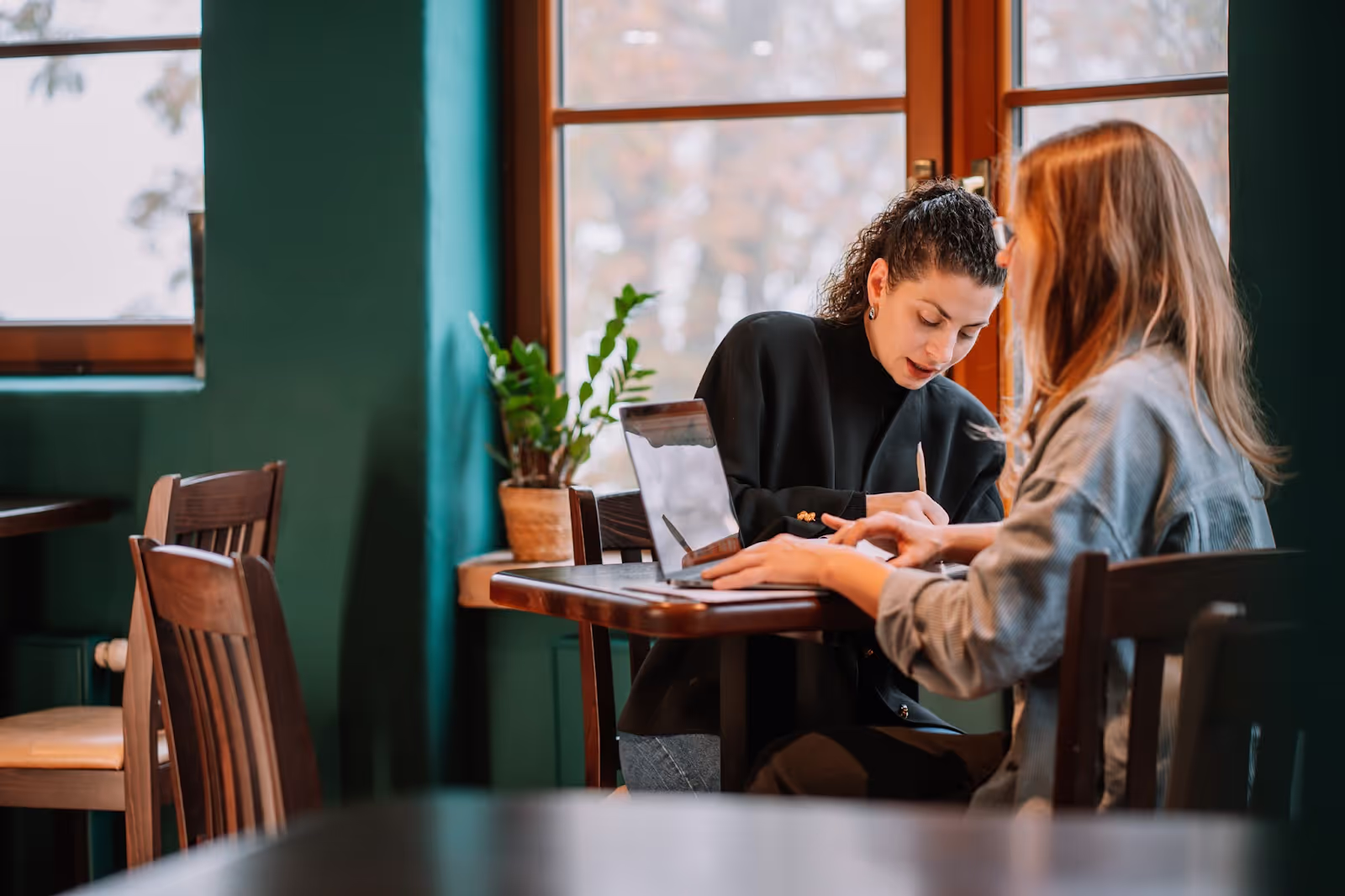 Two people sit at a wooden table, focused on writing, in a room with green walls and large windows. A potted plant decorates the table.