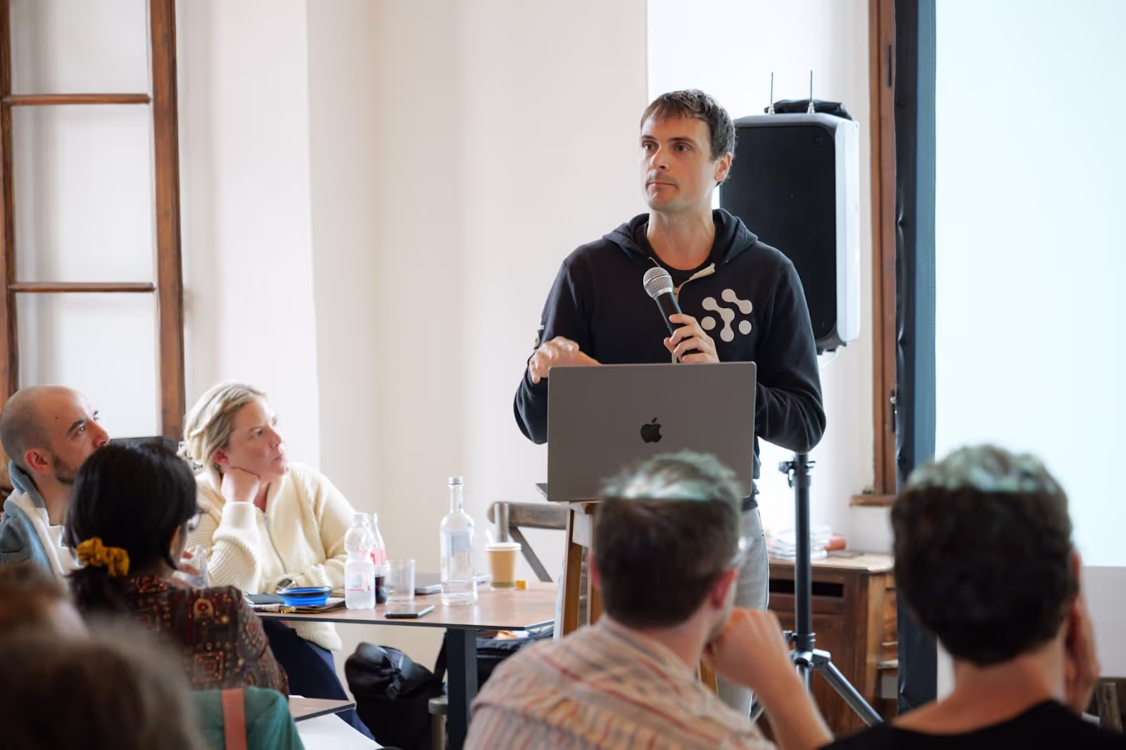 A person holds a microphone, standing at a lectern with a laptop, speaking to an attentive audience seated at tables in a room with wooden accents.