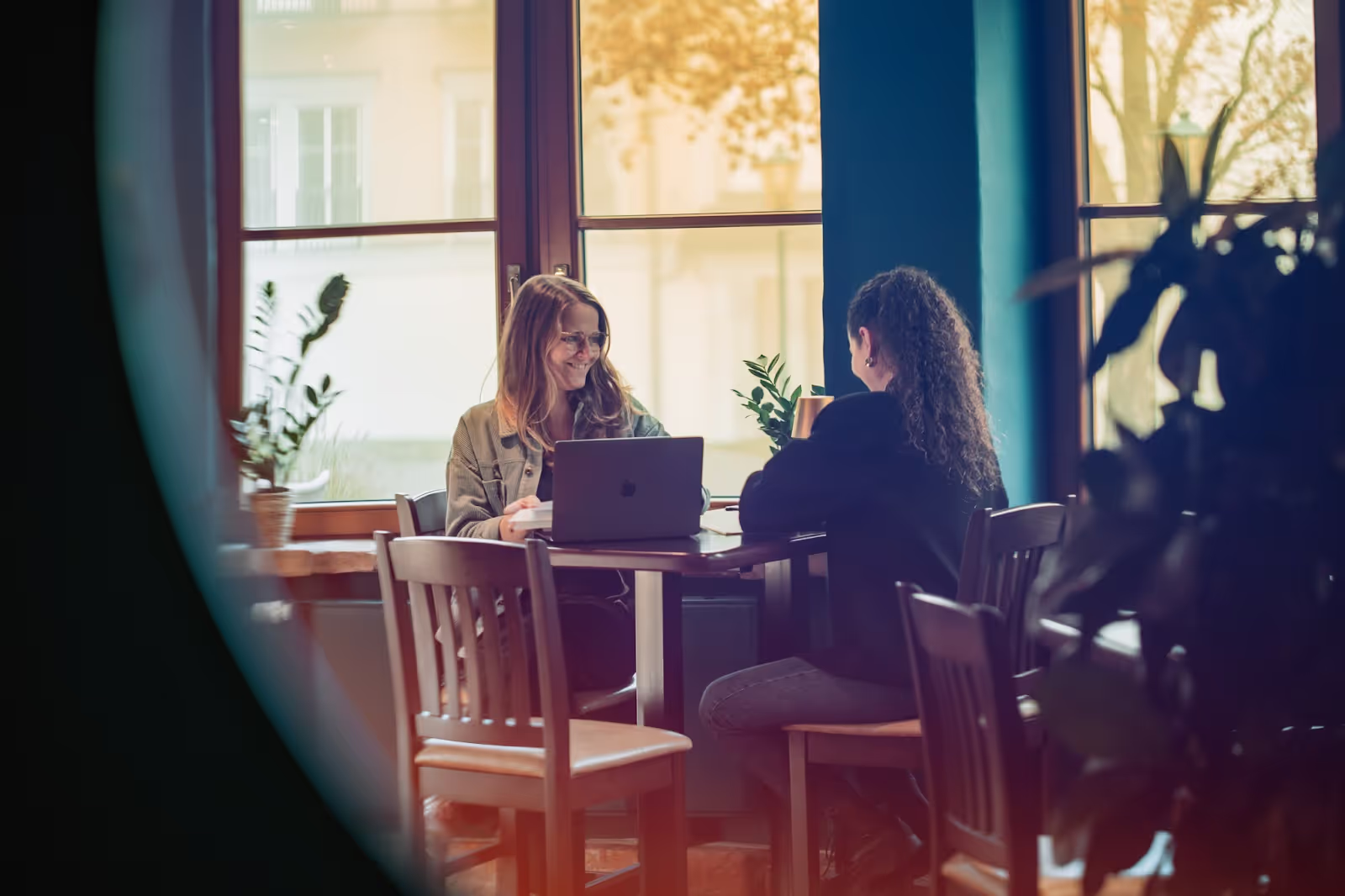 Two people sit at a wooden table, engaged in conversation with a laptop open between them. The setting is a warmly lit room with large windows and potted plants.