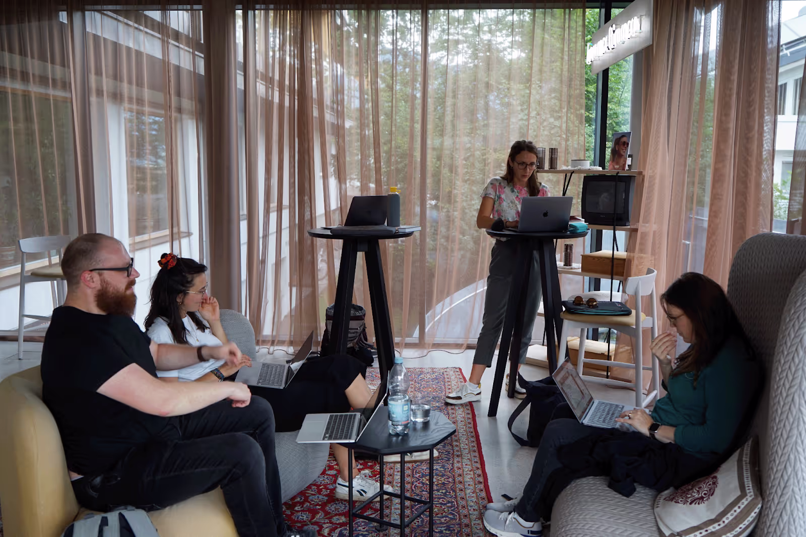 People using laptops, seated and standing, in a cozy room with modern furniture and sheer curtains. A television and shelf with items are in the background.